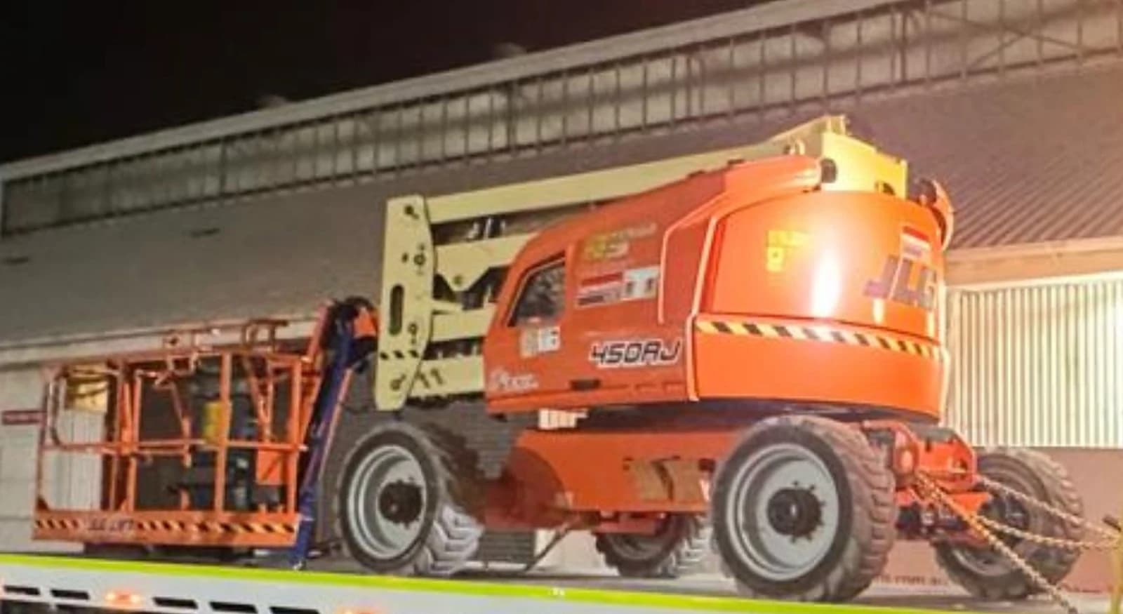 Orange JLG articulating boom lift chained down on a transport deck at night