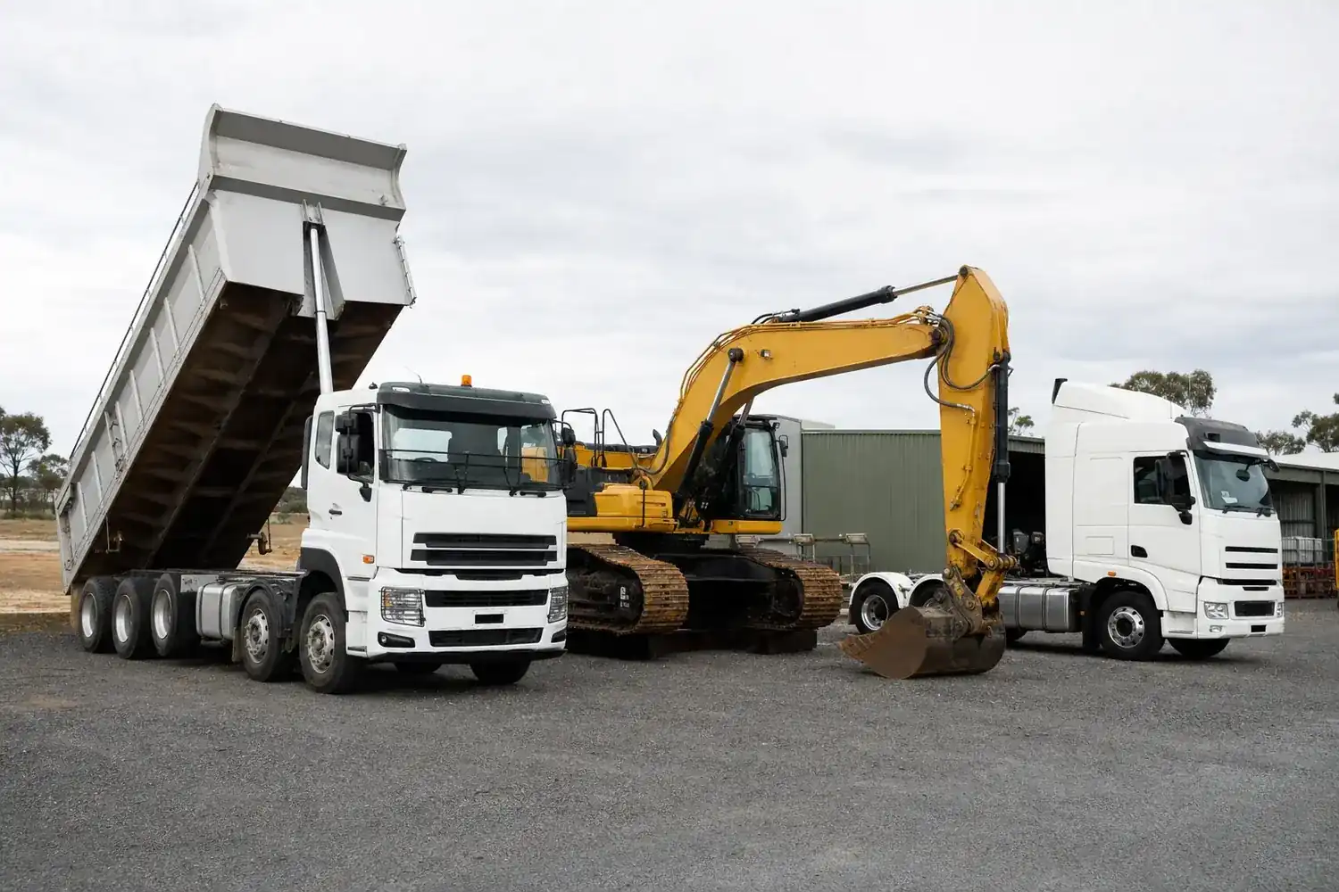 Mixed commercial fleet in an Australian yard with a white tipper truck, tracked excavator and white prime mover parked together