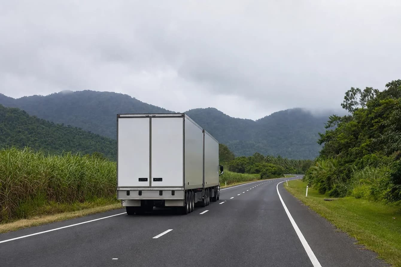 B-double keeping left on a coastal Queensland highway, sugar cane on the verge and misted rainforest ranges ahead