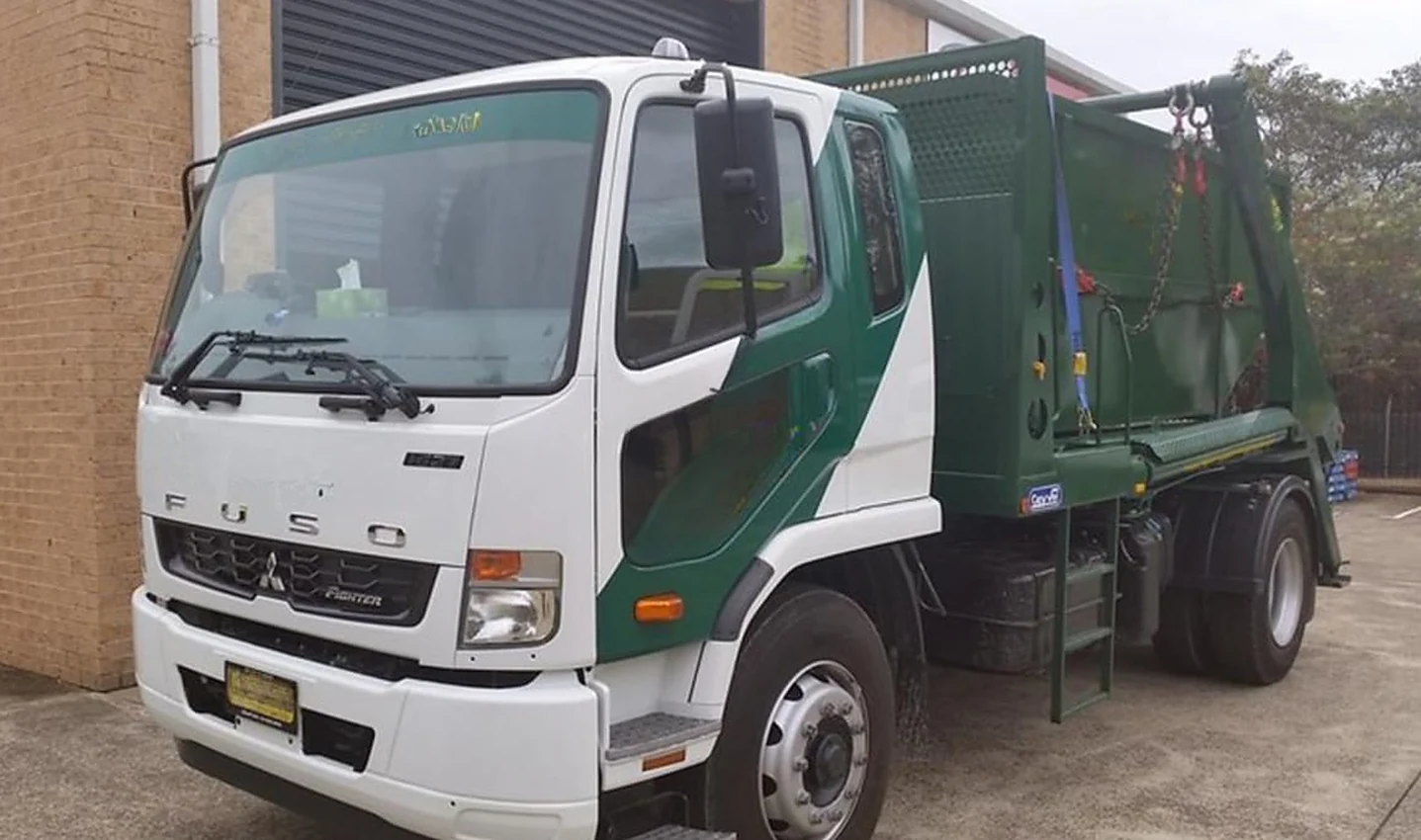 Green and white Australian skip bin truck loaded with a steel skip, lifting chains hooked on, parked beside a warehouse