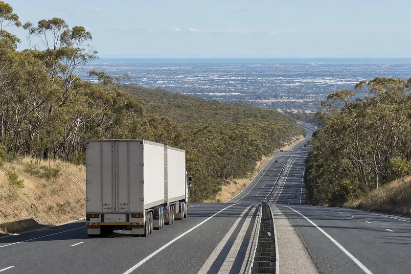 B-double keeping left on a divided freeway descending out of forested hills toward a flat plain in South Australia
