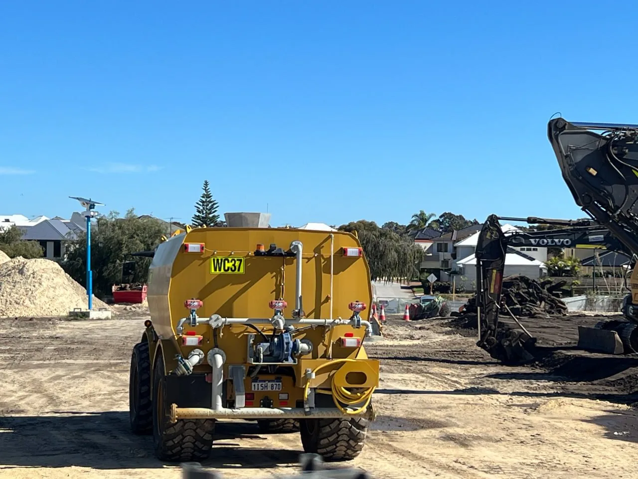 Yellow site water cart with its rear spray bar, parked on a graded earthworks pad